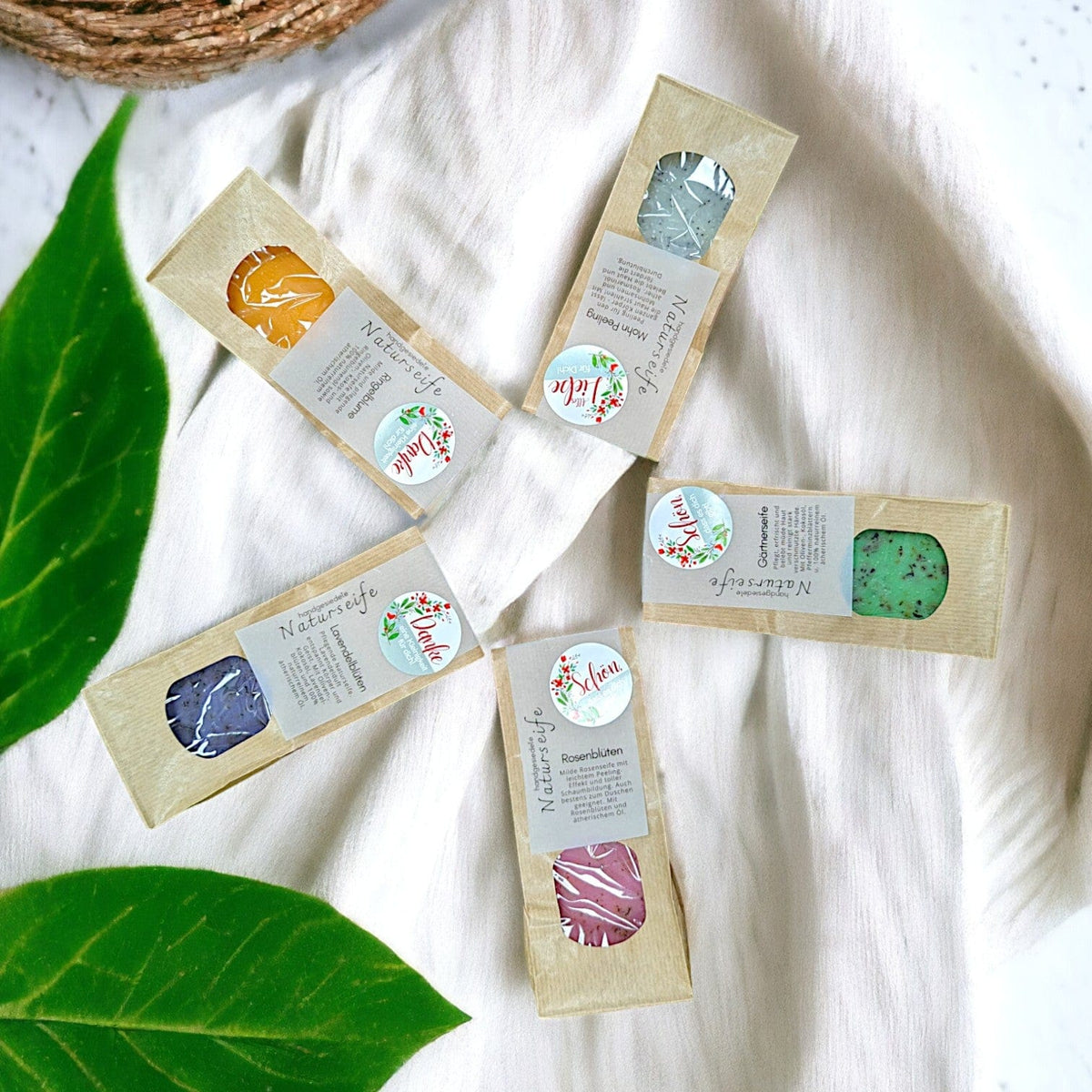 Fünf handgesiedete Naturseifen in Kraftpapierboxen, verschiedenfarbig (blau, grün, rosa, orange), auf weißem Leinen mit…