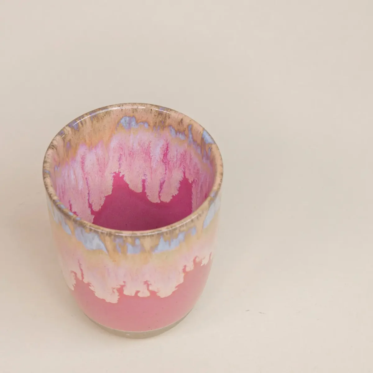 Blick von oben in eine rosafarbene Keramiktasse mit fließender Glasur in Pink, Creme und Hellblau, auf beigem Untergrund.