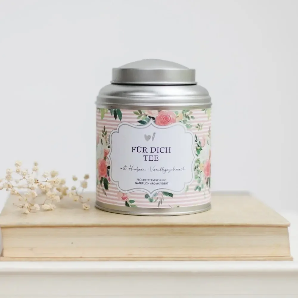 Früchtetee Himbeer Vanille in silberner Dose mit Blumenetikett, neben aufgeschlagenem Buch und Trockenblüten