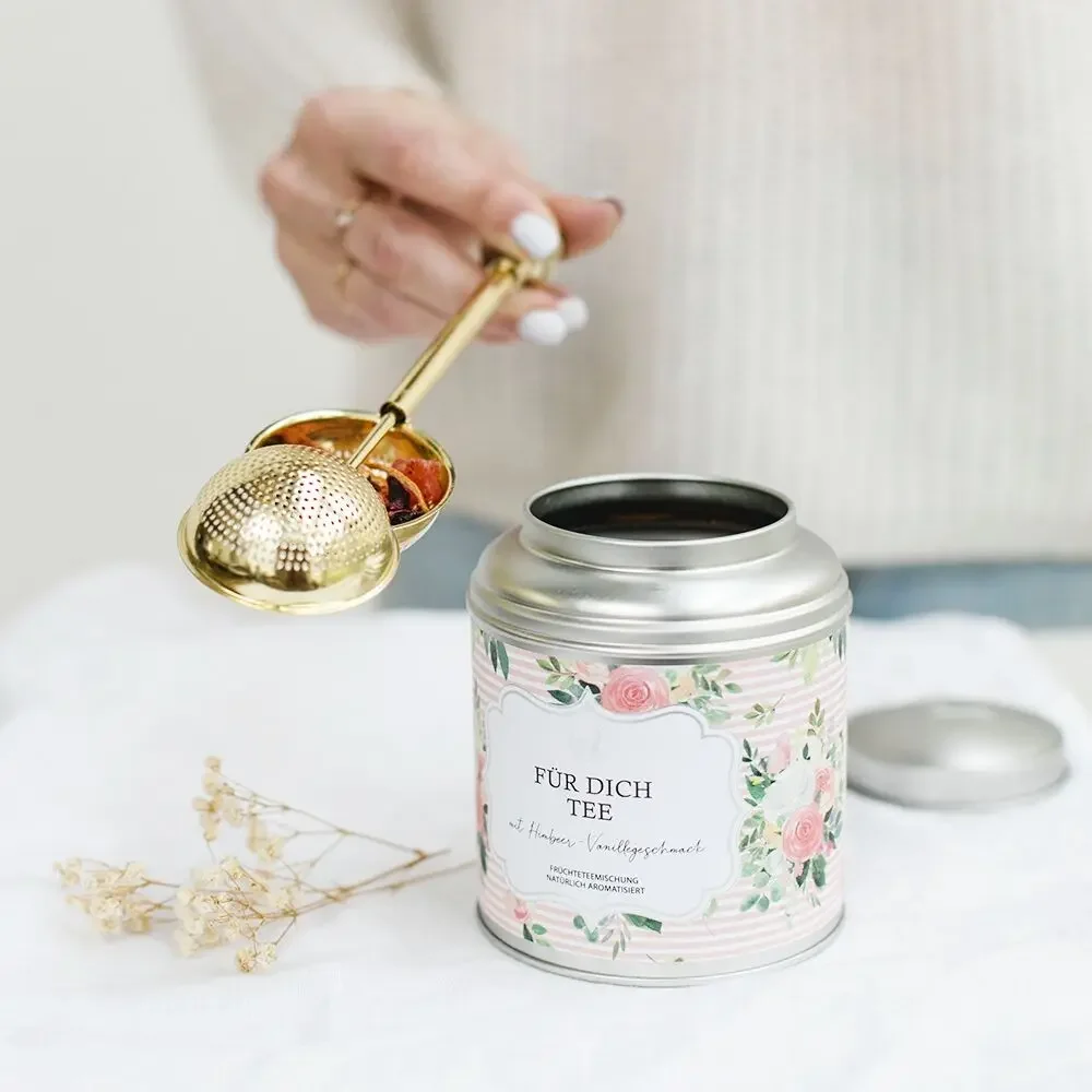 Teekanne mit goldenem Sieb wird über eine Blechdose mit Blumenmotiv gehalten, daneben getrocknete Blüten auf weißem…