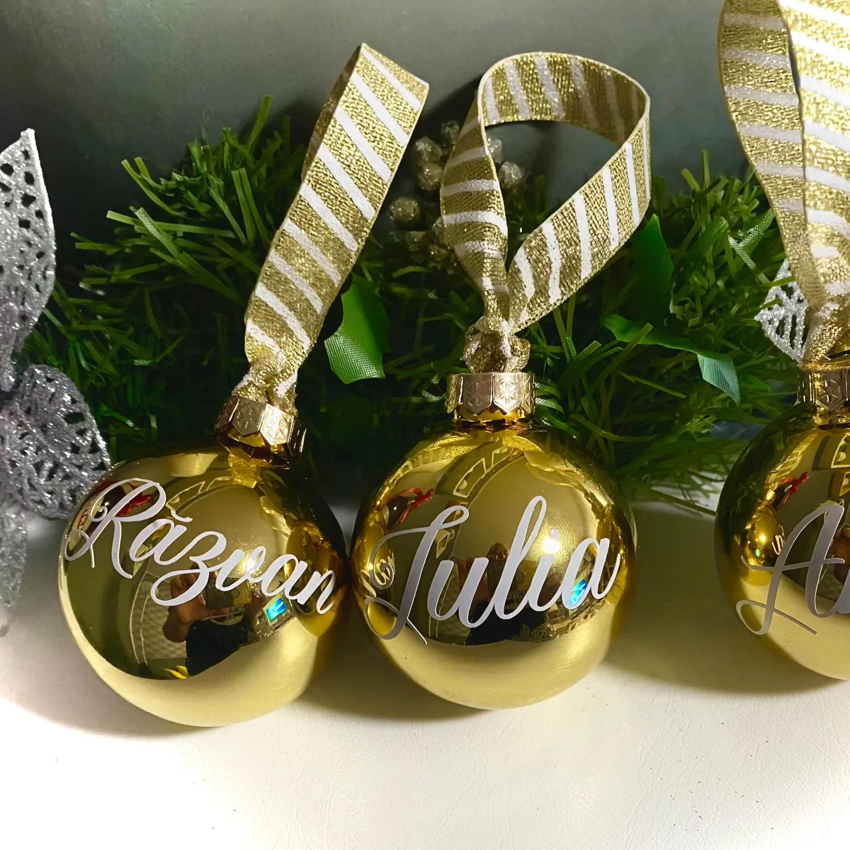 Drei goldglänzende Glaskugeln mit weißen Schriftzügen, links „Răzvan“, mitte „Iulia“, rechts angeschnitten, an gold-weiß…
