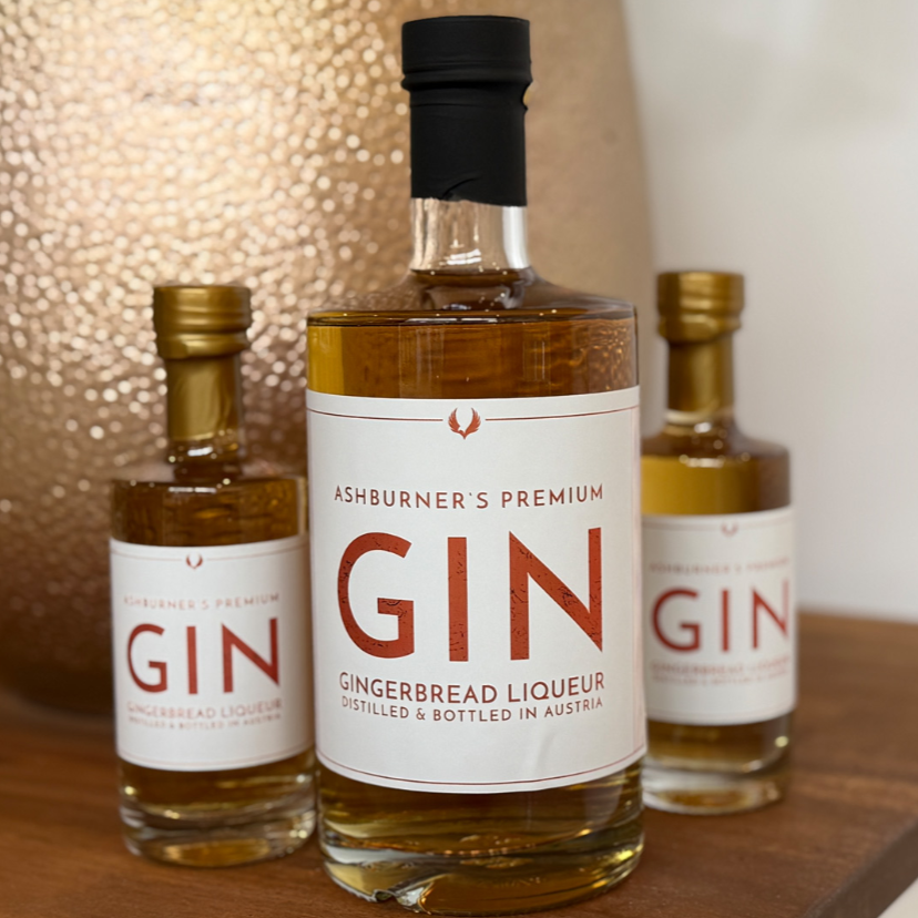 Drei Flaschen Ashburner's Gingerbread Gin Liqueur in unterschiedlichen Größen, Mittelflasche im Fokus mit bernsteinfarbener…