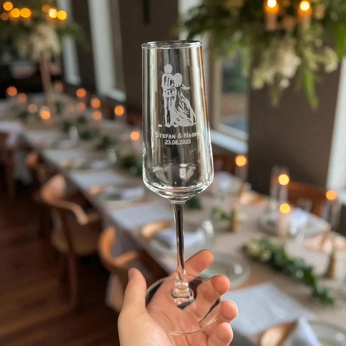 Sektglas mit Gravur in einer Hand, zeigt Namen und Hochzeitsdatum, im Hintergrund festlich gedeckter Tisch mit Kerzen und…