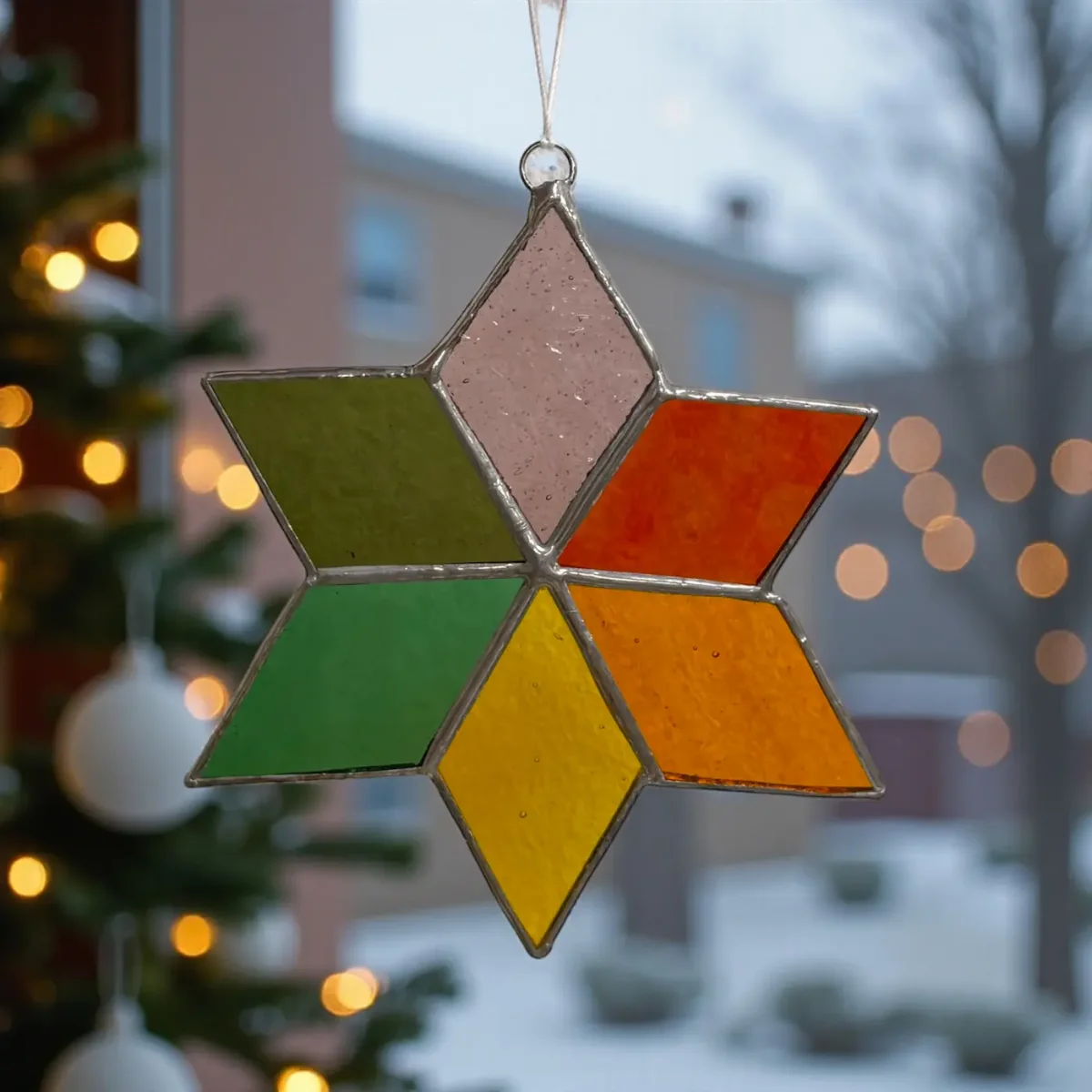 Fensterstern aus Tiffany-Glas, sechszackig, in Grün, Rot, Gelb, Orange und Rosa, hängend vor Fenster mit Weihnachtslichtern
