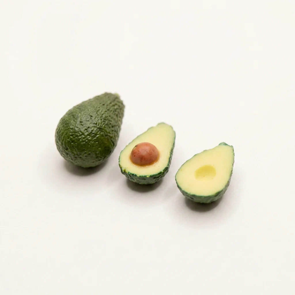 Miniaturavocado aus Polymer Clay: ganze Frucht mit strukturierter grüner Schale, daneben zwei Hälften mit cremefarbenem…