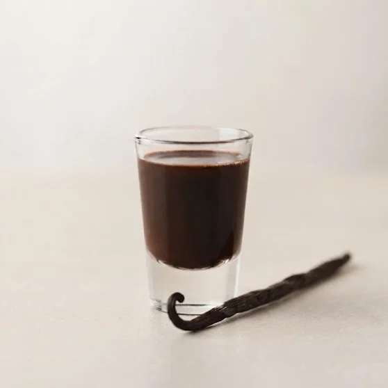 Schuss-Glas mit dunkelbraunem Schokoladenlikör, daneben eine Vanilleschote auf hellem Untergrund