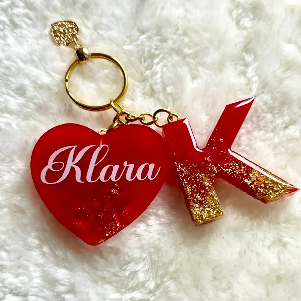 Roter Herz-Schlüsselanhänger mit weißer Schrift „Klara“ und goldener Initiale K aus Resin, auf weißem Fell liegend.