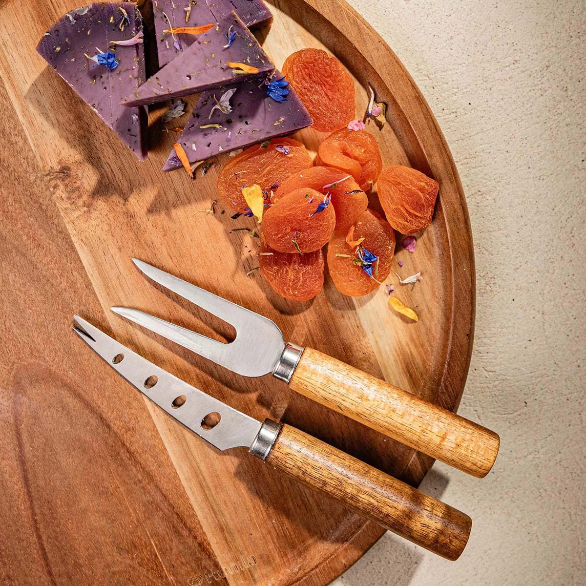 Rundes Holzbrett mit Käse, Schinken und Blüten arrangiert, zwei Edelstahlmesser mit Holzgriffen daneben.