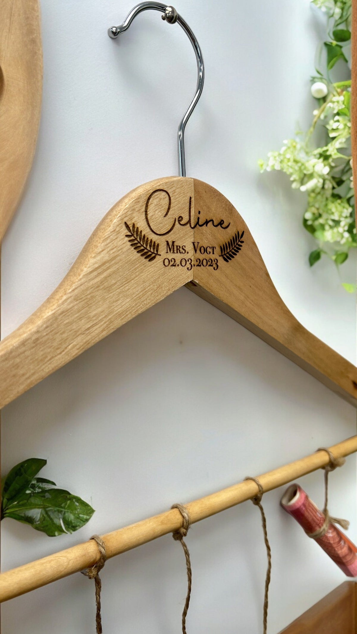 Holzkleiderbügel von vorne, graviert mit Name, Titel und Datum, silberner Haken, an weißer Wand hängend.
