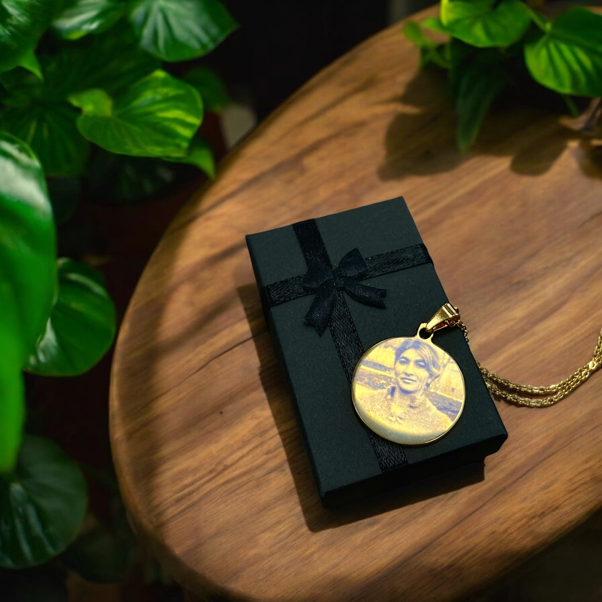 Runder Edelstahl-Anhänger mit Fotogravur auf schwarzer Geschenkbox, an goldener Kette, auf Holzuntergrund mit Pflanzen.
