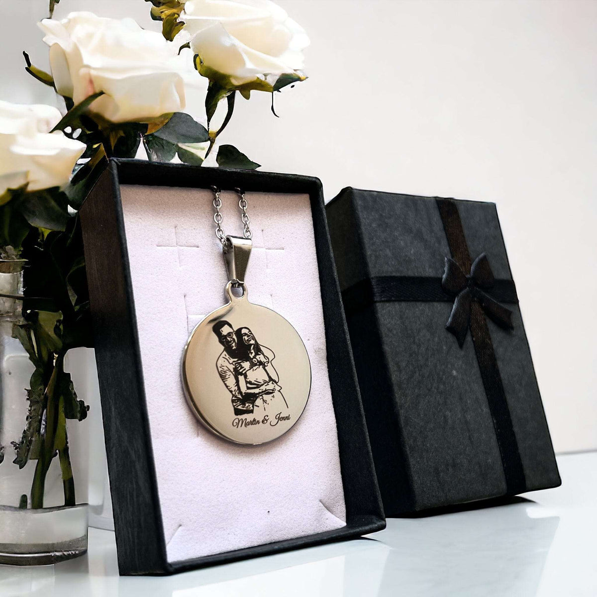 Runde Edelstahl-Halskette mit graviertem Foto in schwarzer Geschenkbox, neben weißen Rosen.