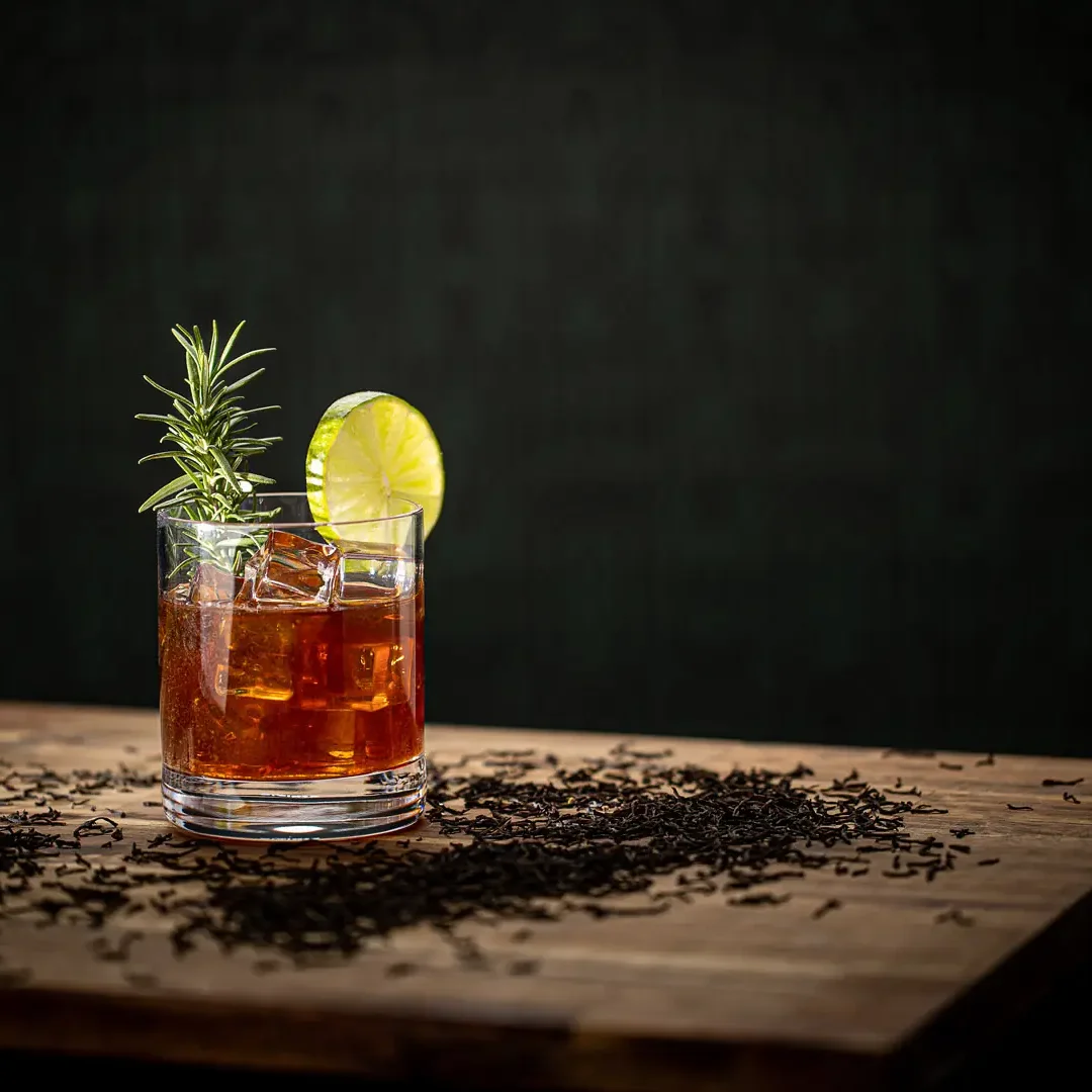 Earl Grey Gin in Glas mit Eis, Zitronenscheibe und Rosmarinzweig, umgeben von Teeblättern auf Holzoberfläche.
