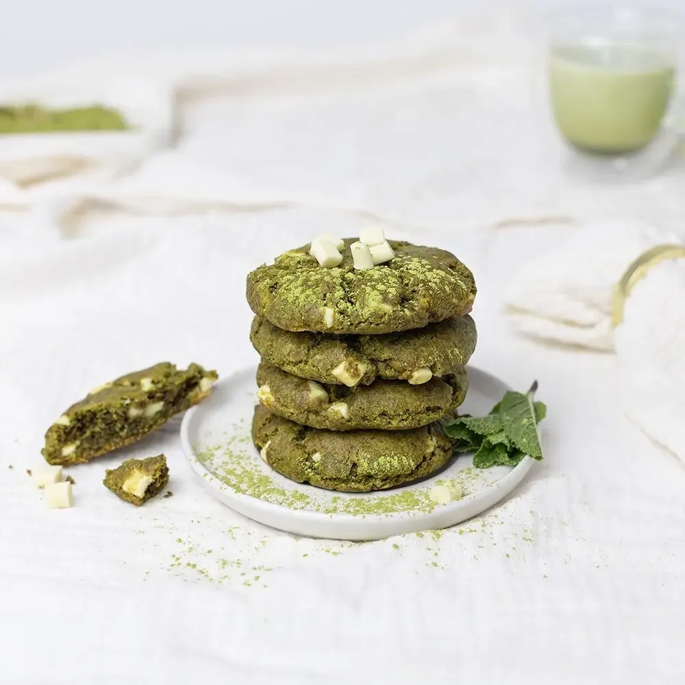 Vier gestapelte Matcha-Cookies mit weißen Schokoladenstücken auf weißem Teller, daneben ein angebrochenes Cookie und grünes…