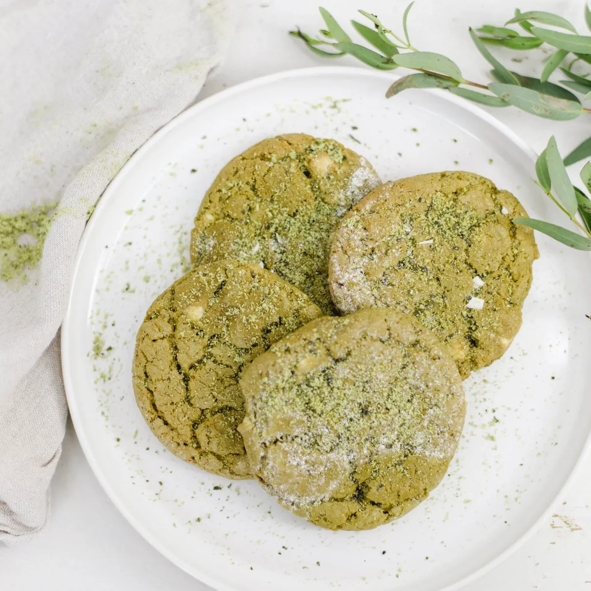 Drei grüne Matcha-Cookies mit Puderzucker auf weißem Teller, daneben frische Matcha-Blätter.