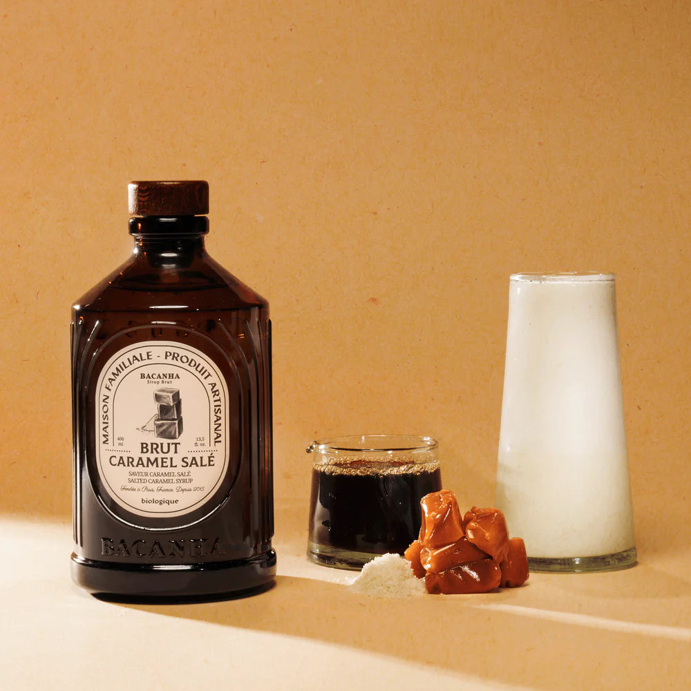 Dunkle Glasflasche mit Bacanha-Etikett neben einem Glas mit dunklem Sirup, Milchglas und Salzkaramell-Bonbons auf beigem…