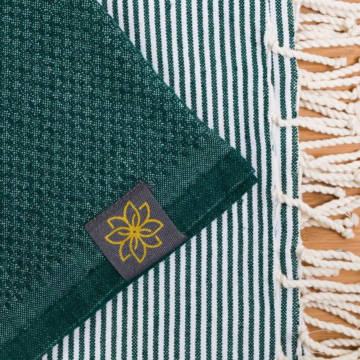 Grünes Fouta-Tuch mit Wabenmuster und weißen Streifen, Detail des eingewebten Jasmin-Logos auf dunklem Label, weiße Fransen…